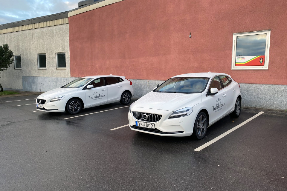 Flen trafikskolas vita bilar mot en tegelvägg.